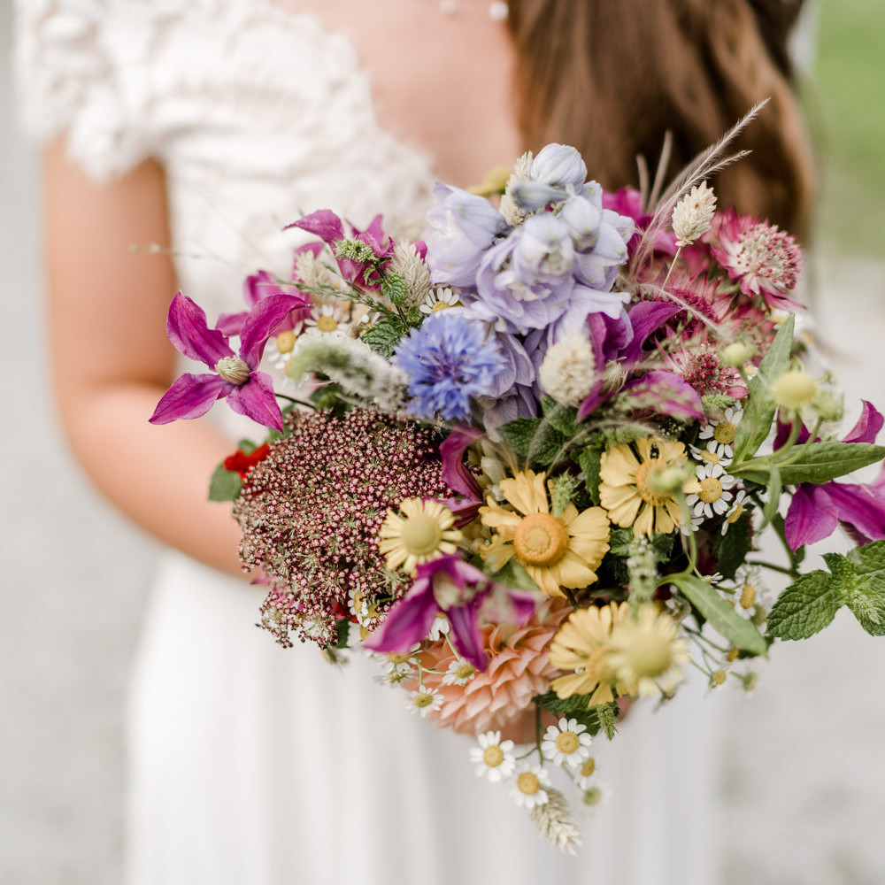 Braut hält bunten Wiesenblumenstrauß von Ganz Unverblümt in die Kamera