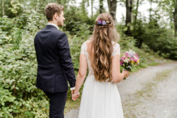Braut und Bräutigam gehen im Wald Hand in Hand mit Strauß und Haardeko von Ganz Unverblümt