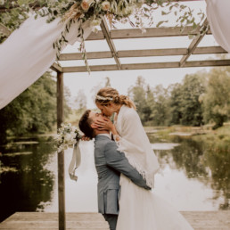 Braut und Bräutigam küssen sich unter Blumenschmuck von Ganz Unverblümt