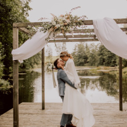 Braut und Bräutigam küssen sich vor See mit Blumenschmuck von Ganz Unverblümt