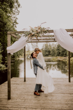 Braut und Bräutigam küssen sich vor See mit Blumenschmuck von Ganz Unverblümt