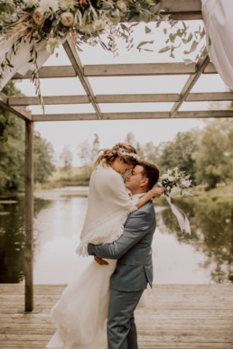 Bräutigam hebt Braut vor Schönsee mit Haarband und Strauß von Ganz Unverblümt