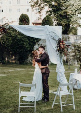 Braut und Bräutigam küssen sich unter Traubogen mit Blumenschmuck von Ganz Unverblümt