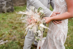 Braut und Bräutigam umarmen sich mit wildem Bohobrautstrauß in zarten Tönen von Ganz Unverblümt