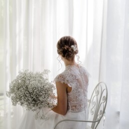 Braut sitzt vor Fenster des Pavillons mit Brautstrauß und Haarschmuck von Ganz Unverblümt