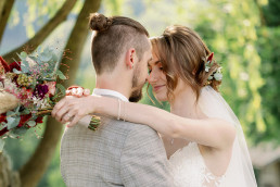 Braut und Bräutigam umarmen sich mit Brautstrauß und Haarblumen von Ganz Unverblümt