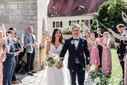Braut und Bräutigam kommen glücklich aus Kirche mit Brautstrauß von Ganz Unverblümt