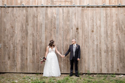 Braut und Bräutigam stehen Händchenhaltetnd vor Scheune mit Strauß von Ganz Unverblümt