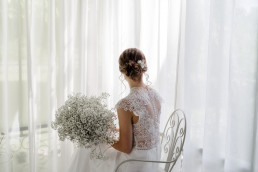 Braut sitzt mit natürlichem Brautstrauß und Haarkranz von Ganz Unverblümt vor Fenster