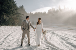 Braut und Bräutigam spazieren Händchen haltend im Schnee mit natürlichem Boho Brautstrauß von Ganz Unverblümt
