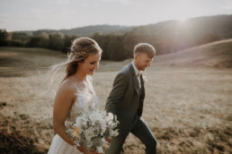 Braut und Bräutigam laufen glücklich mit Brautstrauß von Ganz Unverblümt über Wiese